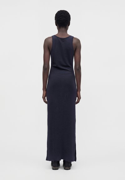 Personne aux cheveux courts et bouclés portant une longue robe sombre sans manches et des chaussures noires, debout de dos sur un fond clair uni.
