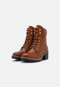 Bottines en cuir marron avec un bout arrondi, des œillets dorés et un laçage. Elles possèdent un talon épais et une semelle en caoutchouc texturée.