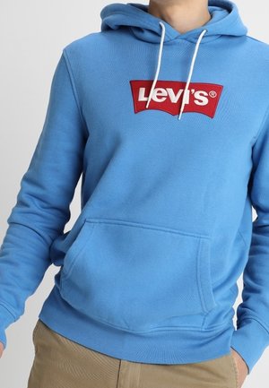 Person trägt einen hellblauen Levi's-Hoodie mit weißen Kordeln und beigen Hosen, steht mit den Händen in der Nähe der Taschen.