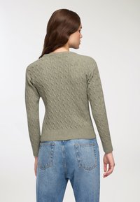 Kobieta z długimi brązowymi włosami ubrana w zielony sweter w warkocz i niebieskie dżinsy, widoczna z tyłu na tle jednolitego białego tła.