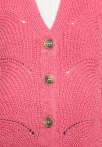 Rosa stickad kofta med V-ringning, fyra runda beige knappar och texturerad sömnad med subtila utskärningar.