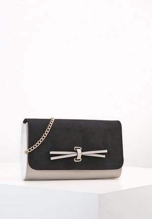 Sort og beige clutch-tasken med en guldkæde som rem og en dekorativ sløjfe foran, vist på en hvid piedestal.