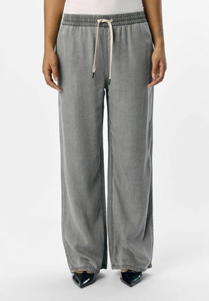 Pantalon gris ample avec taille élastique et cordon beige, porté avec des chaussures noires pointues et un haut blanc sur une personne debout.