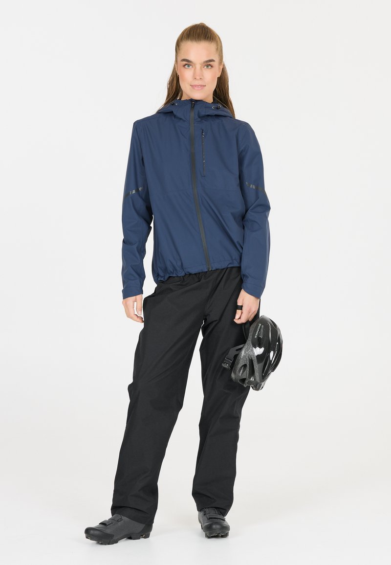 Laivaston vedenpitävä takki, jossa on etuvetoketju ja huppu, yhdistettynä mustiin housuihin ja mustaan pyöräilykypärään, esittelee tyylikästä ja toiminnallista muotoilua.