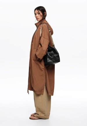 Mujer vistiendo un abrigo largo marrón, pantalones anchos beige y sandalias, llevando un bolso de hombro de cuero negro, de pie contra un fondo blanco.