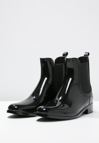 Bottines noires imperméables avec une finition brillante, panneaux latéraux élastiques et petites languettes arrière ; semelle plate et design à bout arrondi.