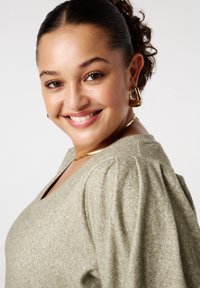 Lichtgroene, gestructureerde trui met een V-hals en pofmouwen. Gouden sieradenaccenten aanvullen de outfit. Schone, neutrale achtergrond.