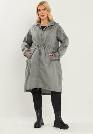 Frau steht und trägt eine lange graue Kapuzenjacke mit Fronttaschen, schwarze Leggings und schwarze Stiefeletten vor einem einfarbigen Hintergrund.