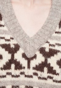 Pull en tricot avec un col en V, fabriqué en laine marron et crème, avec des motifs géométriques complexes. Apparence texturée et chaude.