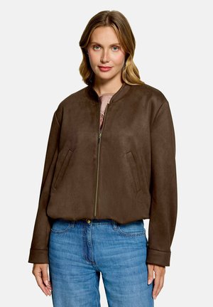 Kvinde med blond hår iført en mørkebrun lynlåstrøje og blå jeans mod en ensartet lys baggrund.