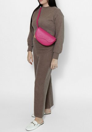 Donna che indossa una maglia marrone a maniche lunghe e pantaloni, scarpe slip-on bianche e una borsa a tracolla rosa acceso.