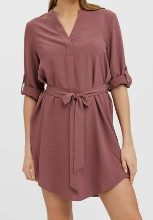 Femme portant une robe mauve ceinturée avec des manches retroussées et un col en V, debout devant un fond clair uni.