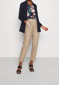 Blazer marine, haut à motif floral, pantalon fuselé en similicuir beige, sandales à talons noirs à bout ouvert. Textures lisses, coupe ajustée.
