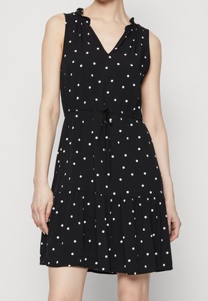 Mouwloze zwarte jurk met witte stippen, V-hals, ceintuur met strik en gerimpelde zoom, gedragen door een staand persoon tegen een effen achtergrond.