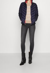Person som bär en marinblå quiltad jacka med röd förstärkning, en beige randig tröja, mörkgrå leggings och svarta höga ankellägenheter.