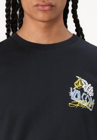 Black cotton t-shirt with crew neck, featuring a colorful graphic print with a yellow emblem and stylized text on the left chest.