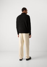 Svart strikket genser med krage, kombinert med kremfargede bukser med sidestriper. Modellen står og viser frem det avslappede designet og passformen.