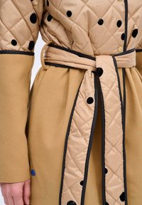 Prošívaná béžová vnější vrstva s černými puntíky, doplněná hladkým hnědým páskem a lemováním. Obsahuje knoflíky a detail s modrým puntíkem.