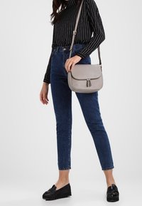 Borsa a spalla in pelle grigia con patta curva, dettagli decorativi in cerniera e tracolla regolabile, abbinata a jeans blu e mocassini neri.