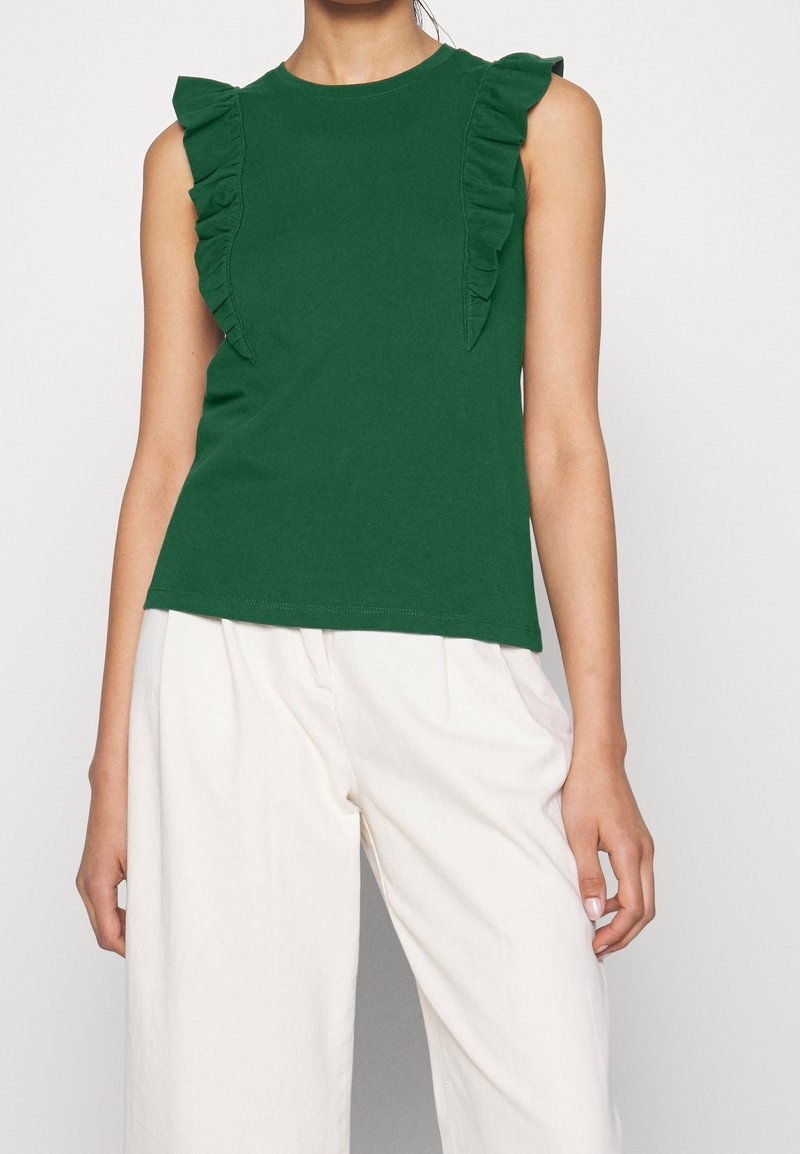 Haut vert sans manches avec des détails froncés le long des épaules, associé à un pantalon blanc ample sur une personne debout.