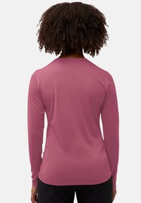 Person mit lockigem Haar, der ein enges, langärmliges, mauvefarbenes Hemd und schwarze Hosen trägt, steht mit dem Rücken zu einem schlichten Hintergrund.