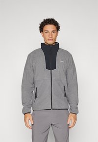 Grå fleecejakke med et svart nylon frontpanel, høy krage, glidelåslukking og to glidelåslommer. Har elastiske mansjetter og snorjustering.