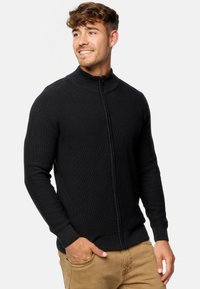 Sudadera negra con cremallera, textura acanalada, cuello alto, diseño ajustado, mangas largas, llevada sobre pantalones beige, acabado de tela suave.