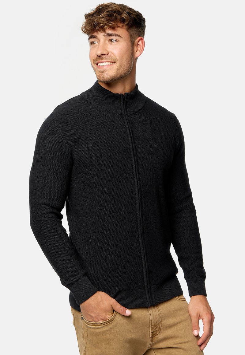 Sudadera negra con cremallera, textura acanalada, cuello alto, diseño ajustado, mangas largas, llevada sobre pantalones beige, acabado de tela suave.