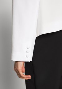Main détendue le long du corps, portant une manche de blazer blanc avec trois boutons recouverts et un pantalon noir sur un fond uni.