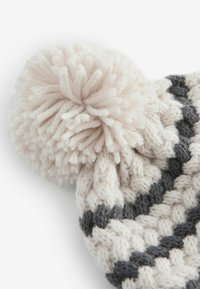 Bonnet en laine tricoté avec un motif beige et gris foncé, surmonté d'un grand pompon beige. Texture douce et forme arrondie.