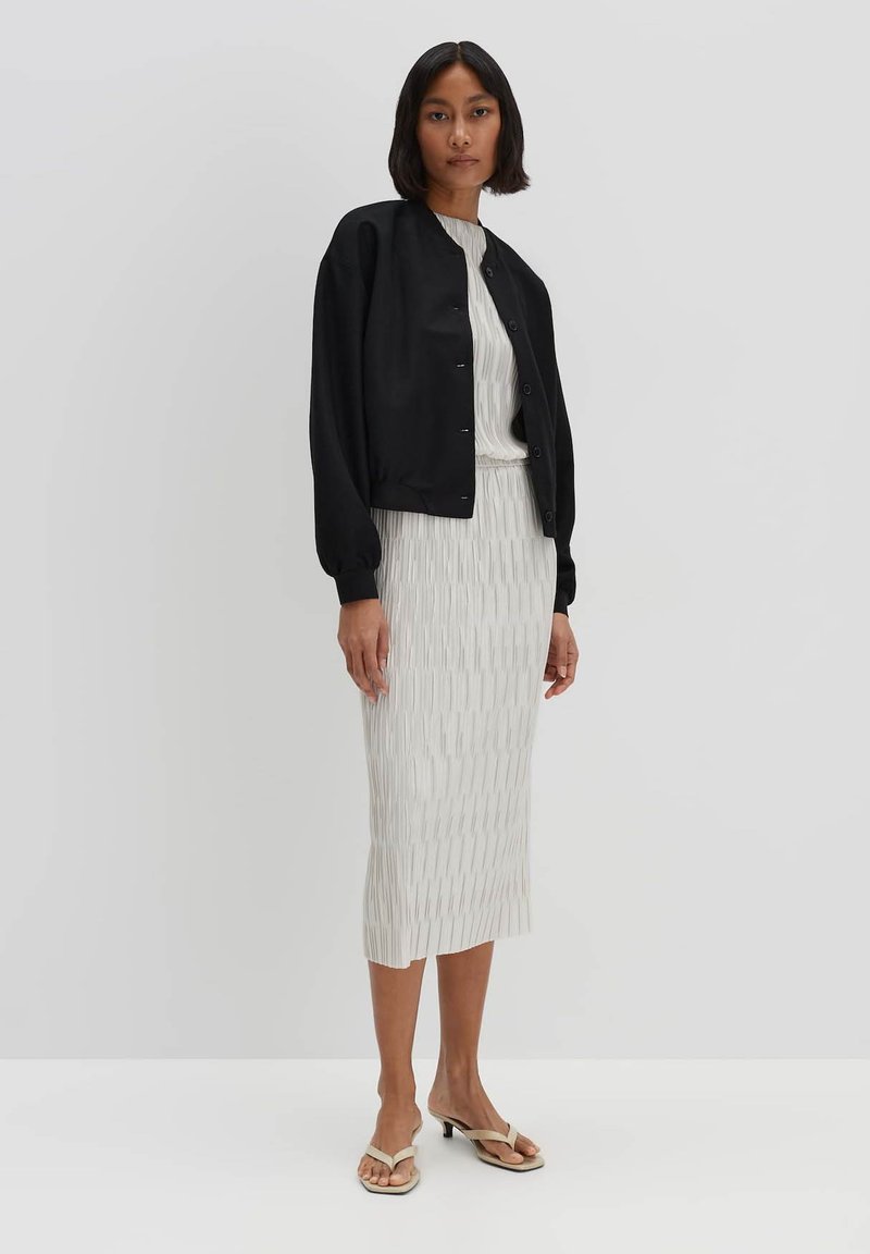 Schwarze Bomberjacke mit Knopfverschluss, kombiniert mit einem ribbeligen, hellgrauen Midikleid mit vertikalen Streifen und einer taillierten Silhouette. Offene Sandalen.