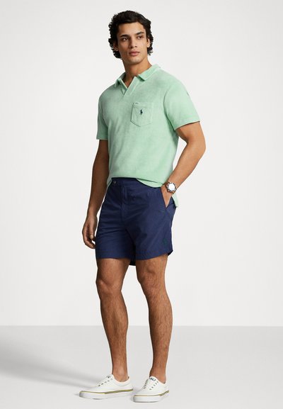 Lichtgroene polo met korte mouwen en zak, gecombineerd met marineblauwe shorts en witte sneakers met gekleurde strepen.