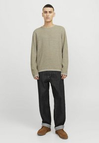 Hellbrauner Strickpullover mit rundem Halsausschnitt und langen Ärmeln, kombiniert mit dunkelblauen Jeans mit umgeschlagenem Saum und braunen Wildlederschuhen.