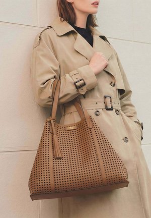 Femme portant un trench-coat beige, transportant un grand sac fourre-tout en cuir marron clair avec des découpes en forme de cœur et un détail à pampilles sur l'épaule.