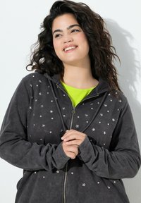 Sudadera con cremallera de color gris oscuro con acentos de estrellas, que presenta una textura suave y un corte relajado. Capa interior de color verde brillante visible por debajo.