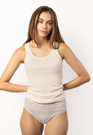 Young woman with long brown hair wearing a white sleeveless top and striped underwear, standing with hands on hips against white background.