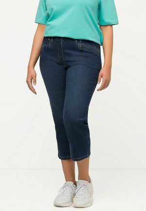 Dunkelblaue Denim-Caprihosen mit engem Schnitt, ausgestattet mit Gesäßtaschen und einem Reißverschluss. Kombiniert mit einem türkisfarbenen kurzärmeligen Top und weißen Turnschuhen.