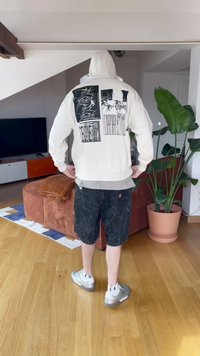 Sudadera blanca con estampados gráficos en la espalda, pantalones cortos negros y zapatillas plateadas. La habitación presenta plantas y un televisor.
