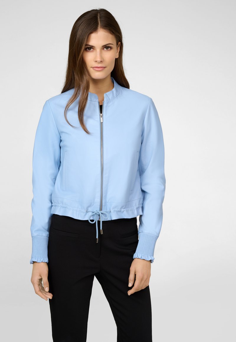 Hellblaues, kurz geschnittenes Jacket mit Reißverschluss vorne, gerippten Bündchen und Kordelzugsaum; glattes Material mit einem subtilen Glanz. Schwarze Hose darunter sichtbar.