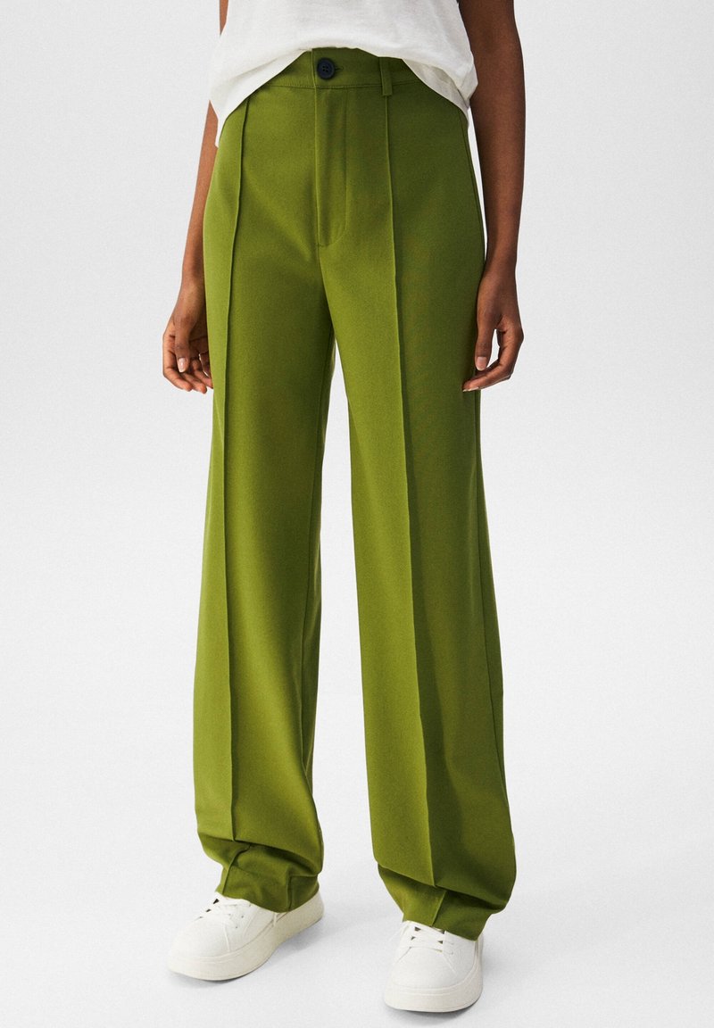 Personne portant un pantalon taille haute, jambes larges, vert olive avec plis à l'avant et des baskets blanches, debout devant un fond uni.
