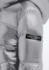 Close-up van een glanzende zilveren pufferjacket-mouw met een zwart etiket "Frieda & Freddies New York" erop genaaid.