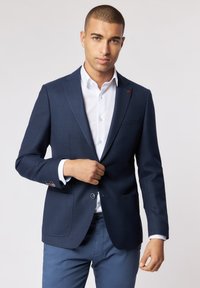 Marineblauer Blazer aus strukturiertem Stoff, mit zwei Vordertaschen und einem roten Akzent am Revers. Kombiniert mit einem weißen Hemd und blauen Hosen.
