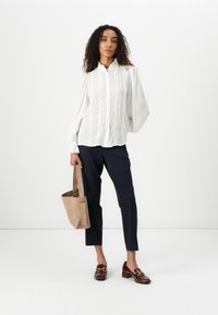 Vit, rynkad blus med vertikala ränder, i kombination med marinblå tapered byxor och bruna loafers; håller en tan mocka tote-väska.