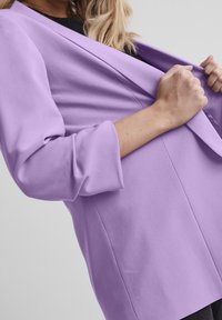 Kvinna som justerar slaget på en ljuslila kavaj med uppvikta ärmar över en svart topp, framför en neutral bakgrund.