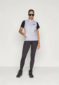 Ljusgrå puffer väst över en svart skjorta, kombinerat med svarta leggings och kraftiga svarta stövlar. Accessoarer inkluderar blåa solglasögon.