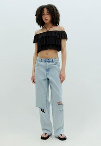 Schwarzes, schulterfreies Croptop mit Rüsche und Spaghettiträgern, kombiniert mit hellblauen, ausgefransten weit geschnittenen Jeans und schwarzen Flip-Flops.