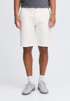 Homme portant un short blanc longueur genoux, des baskets grises, des chaussettes blanches et une chemise grise, debout devant un fond uni.