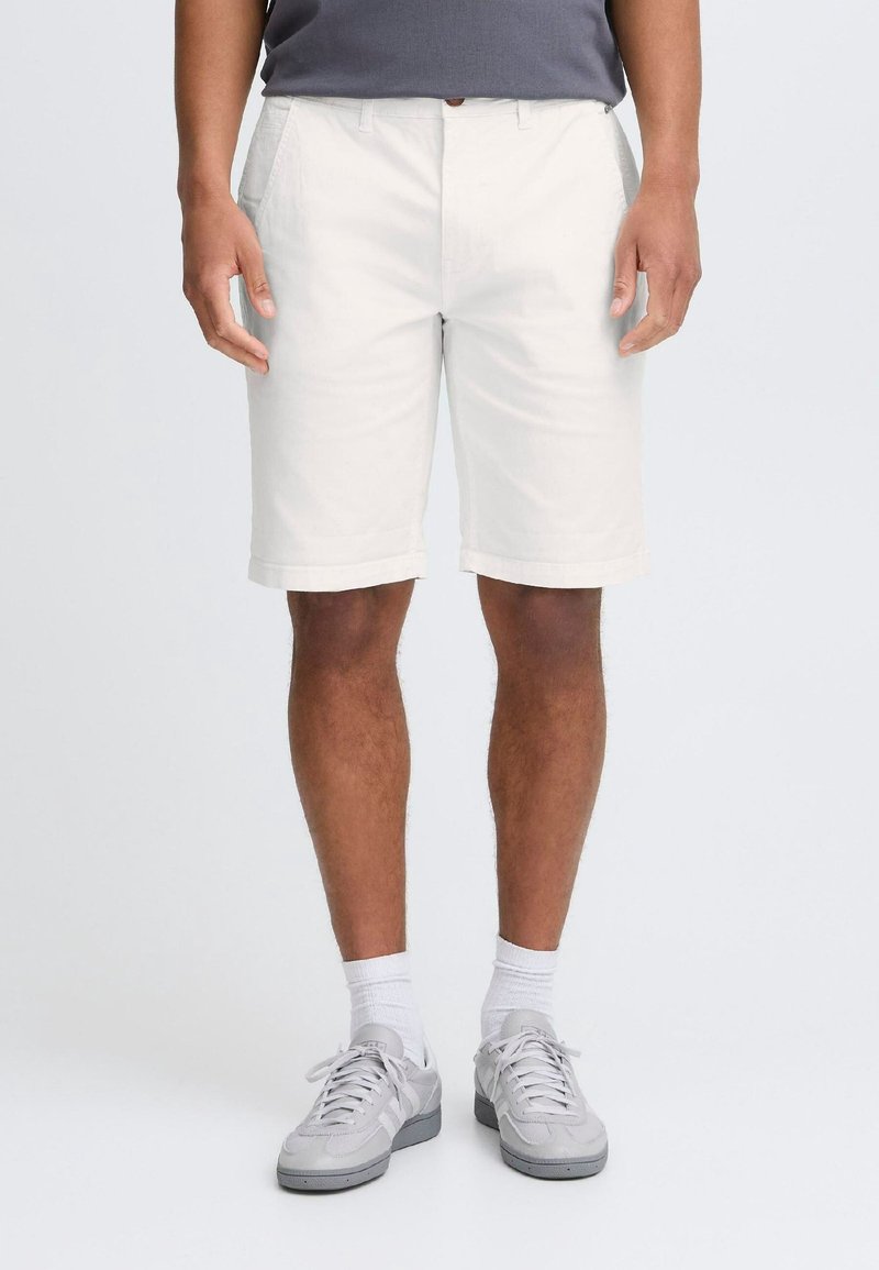 Homme portant un short blanc longueur genoux, des baskets grises, des chaussettes blanches et une chemise grise, debout devant un fond uni.