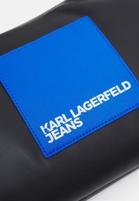 Svart läderpouch med en slät yta, prydd med en ljusblå rektangulär lapp och vit text "KARL LAGERFELD JEANS".