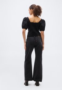 Femme aux cheveux bouclés portant un haut noir à manches bouffantes, un jean large taille haute foncé et des chaussures noires à talons, debout devant un fond blanc.